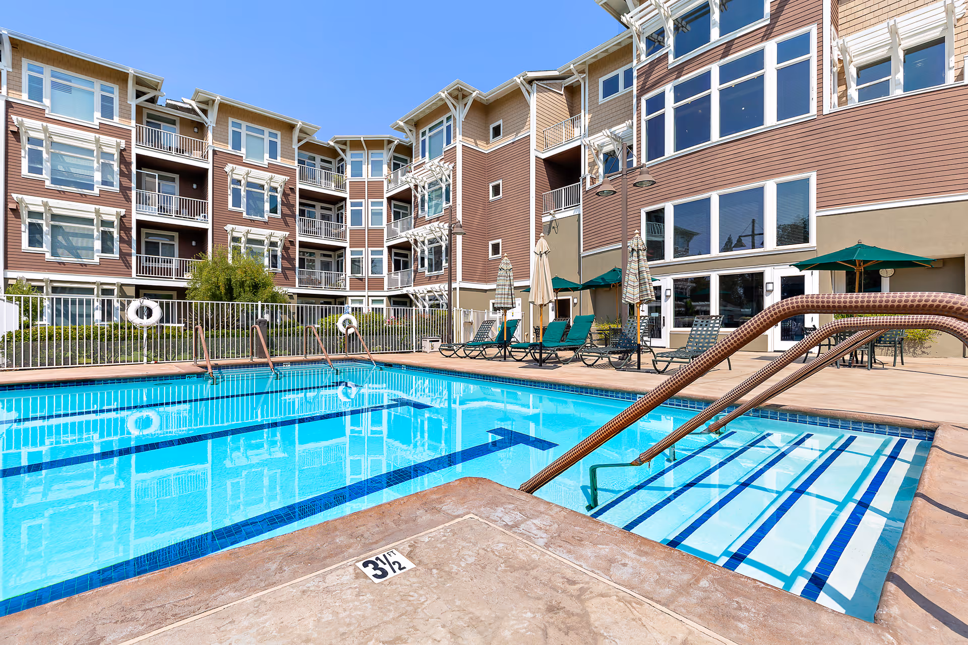 Outdoor swimming pool with clear blue water surrounded by a concrete deck. Several lounge chairs and umbrellas are arranged around the pool. In the background, there is a multi-story residential building with balconies and large windows under a clear blue sky.