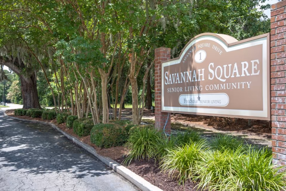 Entrance sign for Savannah Square Senior Living Community surrounded by green trees and bushes along a paved driveway.