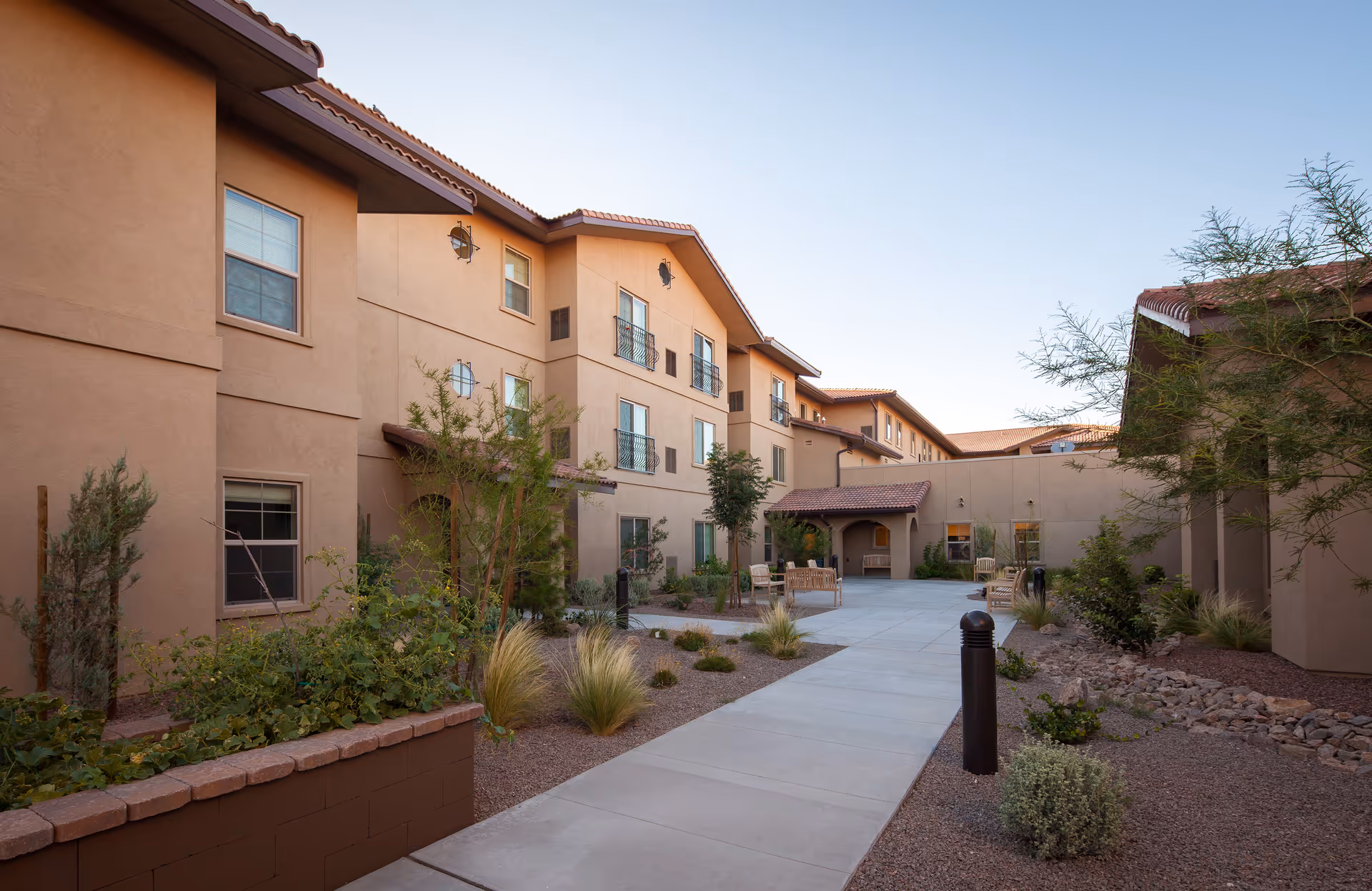 Outdoor courtyard with a paved walkway, seating areas, and landscaping between multi-story tan stucco senior living buildings.