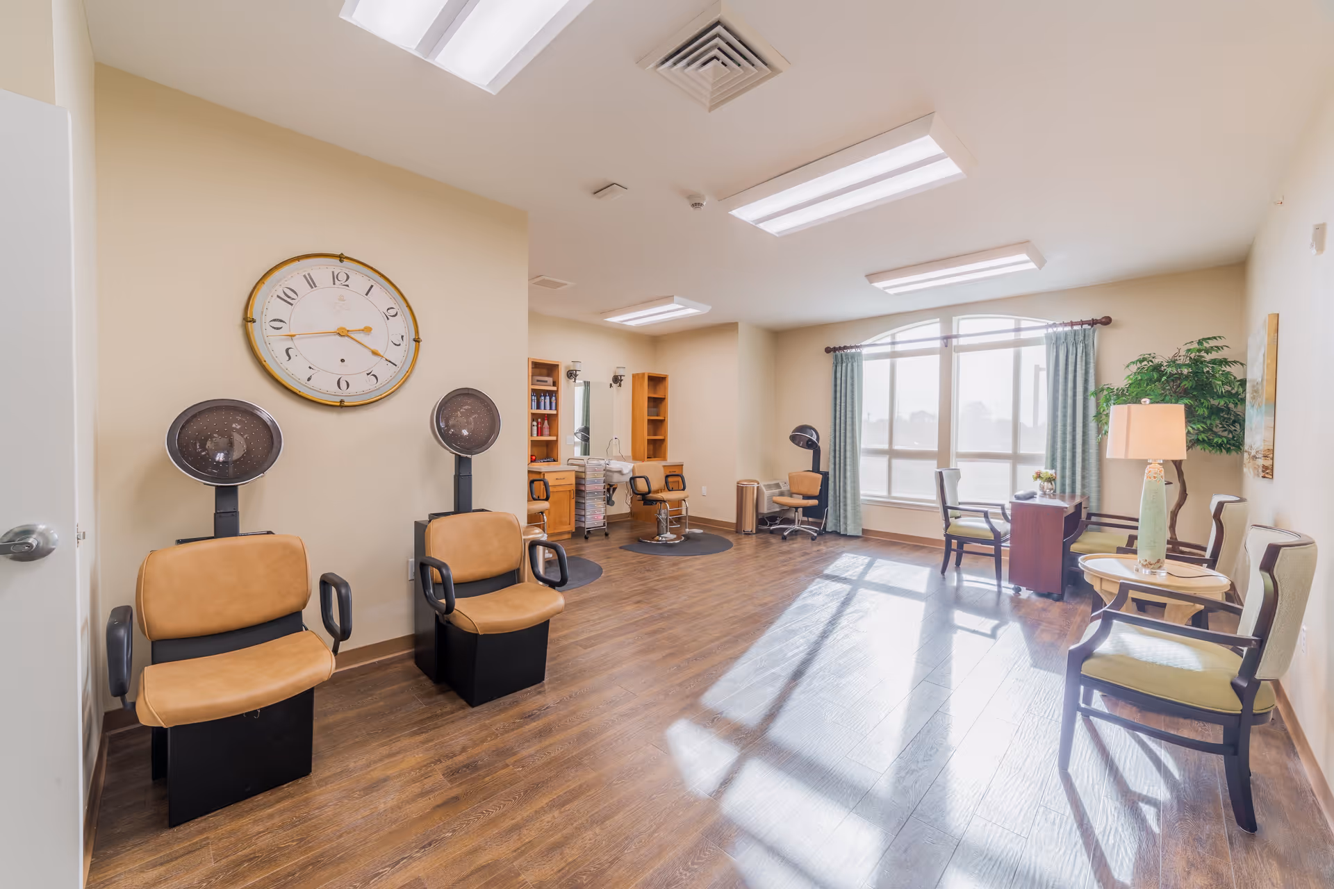 A bright and spacious hair salon area with two vintage-style hair dryers and two salon chairs on the left side. In the background, there are two more salon chairs with mirrors and shelves holding hair products. On the right side, there is a seating area with three chairs, a small round table with a lamp, and a large window with green curtains letting in natural light. The room has wooden flooring and beige walls.