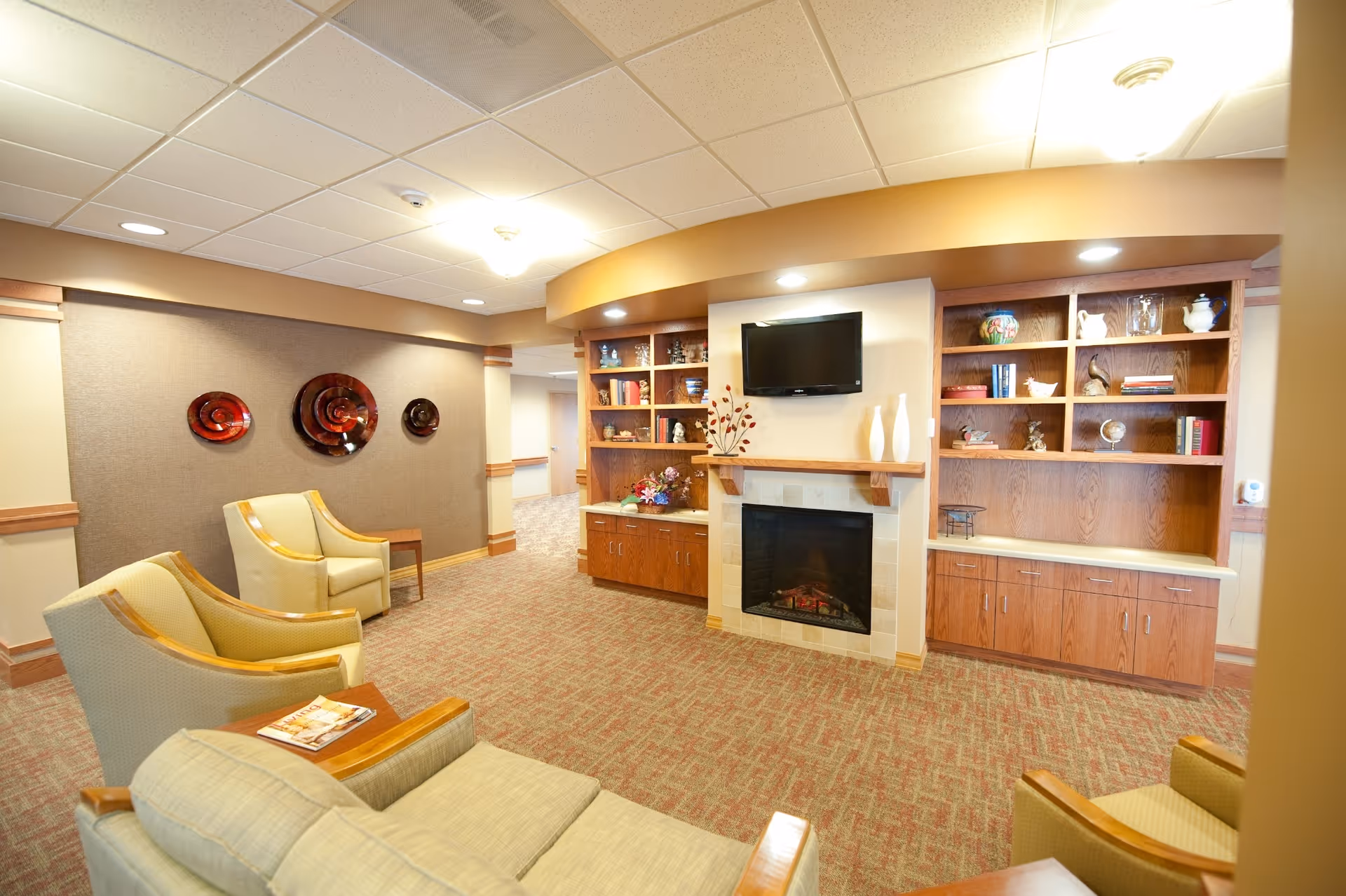 A cozy senior living common room with armchairs, a fireplace, built-in shelving and a wall-mounted TV.