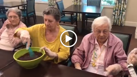 Three elderly women sitting at a table in a dining or activity room. One woman in a yellow shirt is stirring a green bowl with a blue spatula, while the other two women watch. The room has tables and chairs and windows with curtains in the background.