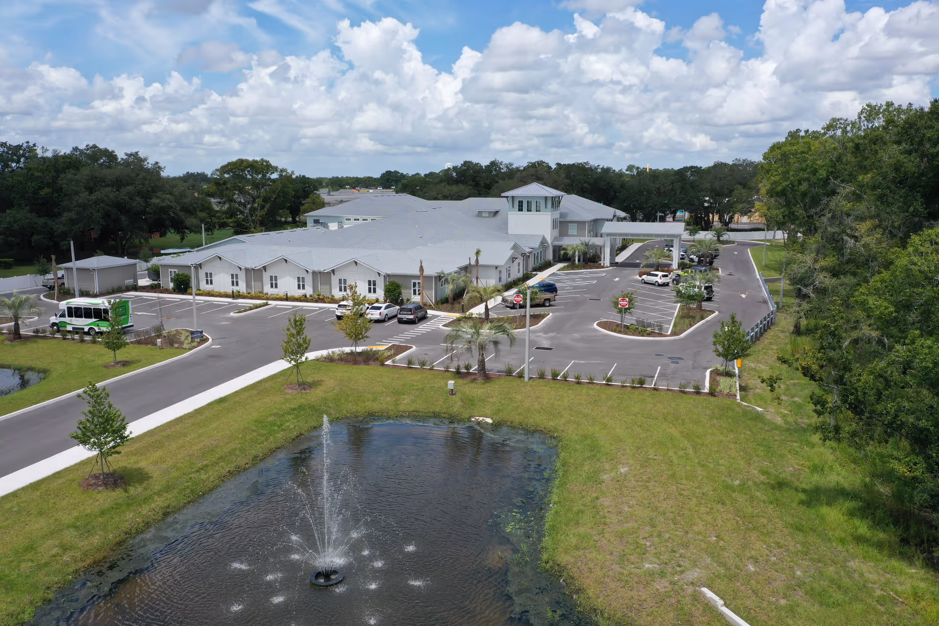 Aerial view of The Canopy at Walden Woods senior living facility showing a large single-story building with a gray roof, surrounded by parking lots with several cars, landscaped greenery, and a pond with a water fountain in the foreground under a partly cloudy sky.