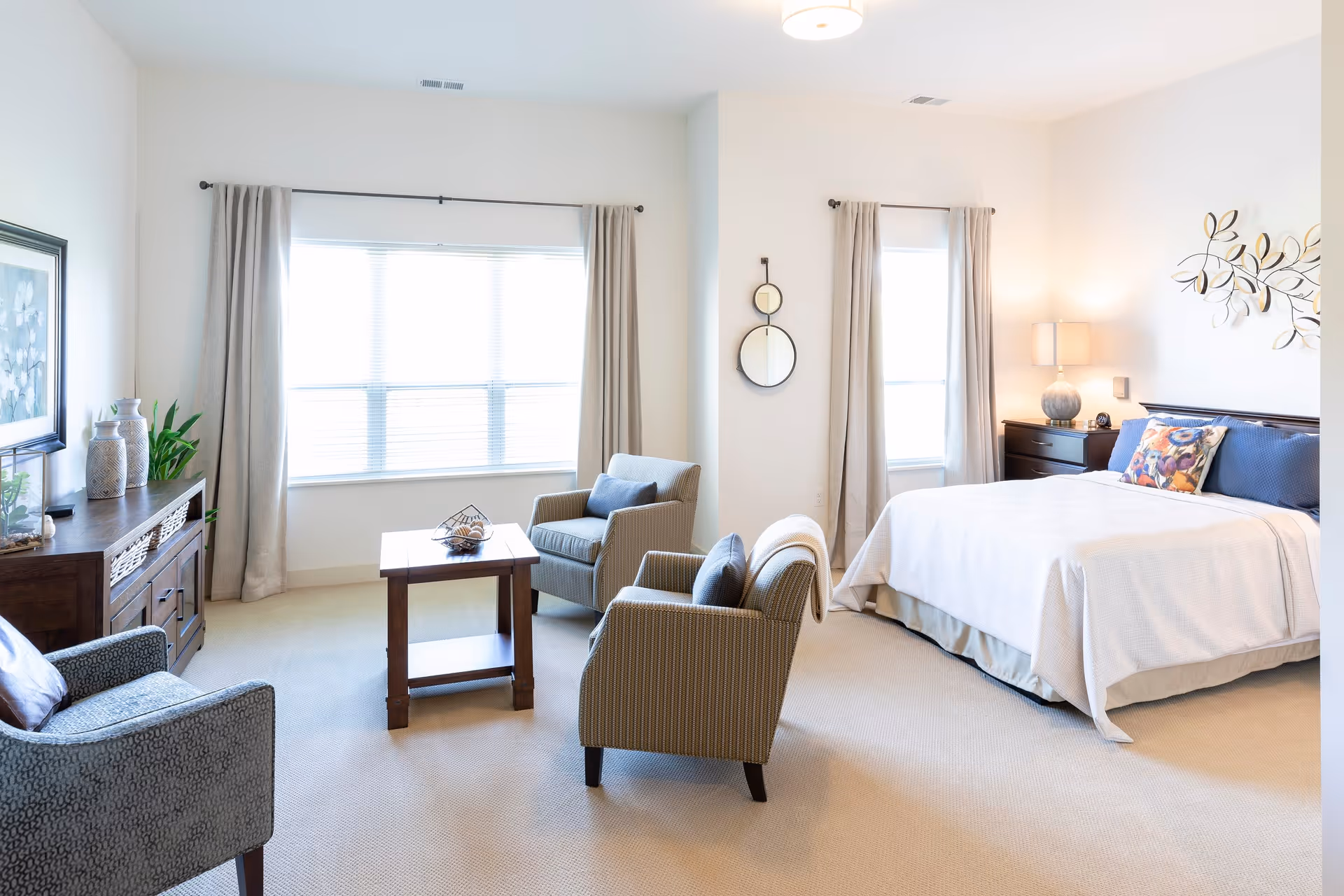 A bright and spacious senior living bedroom featuring a large bed with white bedding and colorful pillows, two armchairs with cushions around a wooden coffee table, a wooden dresser with decorative vases and plants, two windows with beige curtains, a bedside table with a lamp, and wall decor including a metal leaf sculpture and two round mirrors.