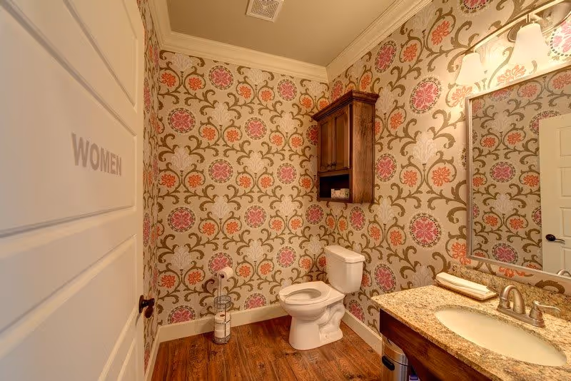 A women's bathroom with floral patterned wallpaper, a white toilet, a wooden cabinet mounted on the wall, a granite countertop with a sink, a mirror above the sink, and wooden flooring.