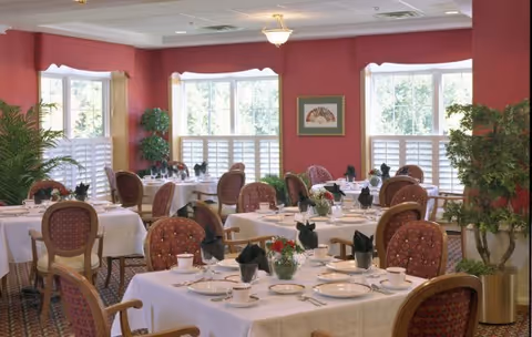 A dining room with multiple round tables covered with white tablecloths, each set with plates, cups, and black folded napkins. The room has red walls, large windows with white shutters, and several potted plants. Upholstered chairs with wooden frames surround the tables.