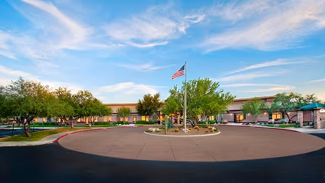 Wide exterior view of The Highlands at Dove Mountain facility showing a circular driveway with an American flag in the center, surrounded by trees and landscaping under a partly cloudy sky.
