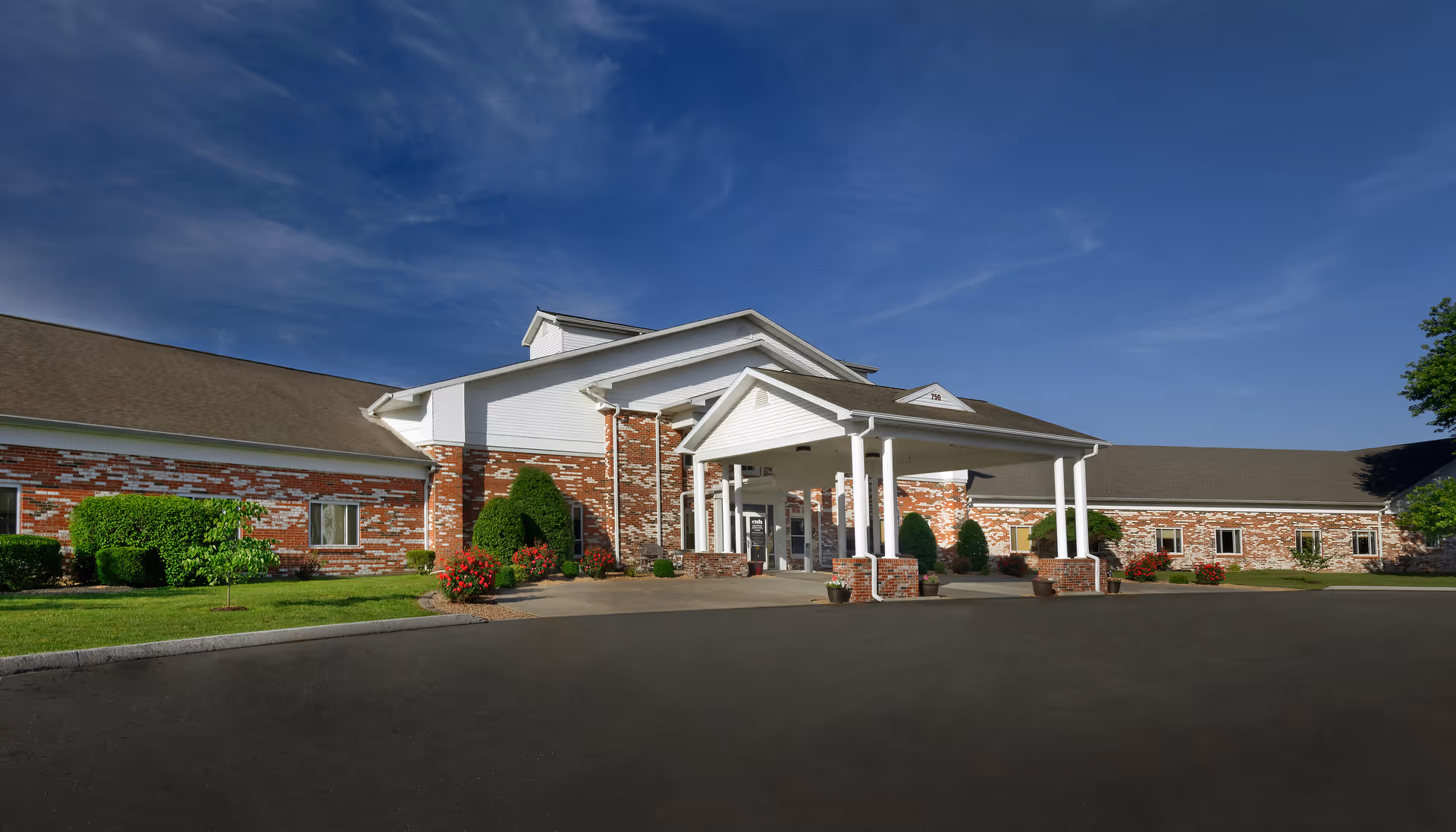 Single-story brick healthcare building with a white columned covered entrance, landscaped lawn, and clear blue sky.