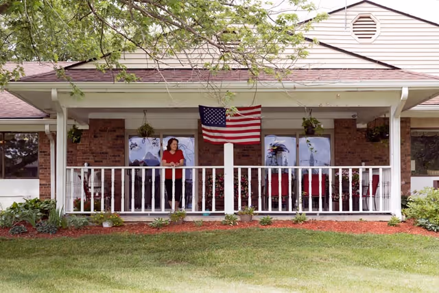Front porch of a brick nursing facility with a white railing, hanging plants, an American flag, and a person standing on the porch overlooking a lawn.