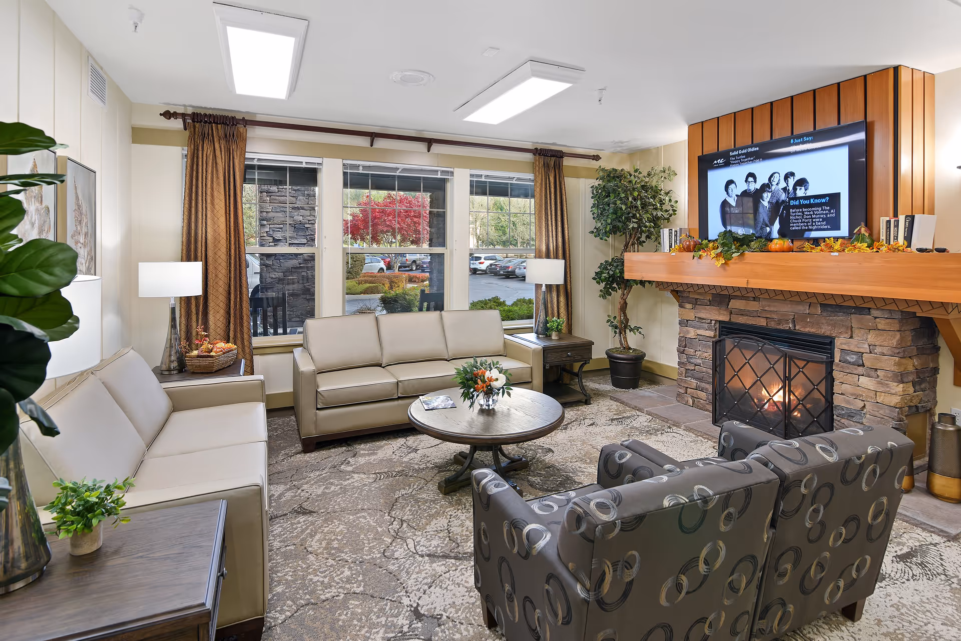 A cozy living room with beige sofas and patterned armchairs around a round coffee table, a stone fireplace with a TV above, and large windows looking outside.