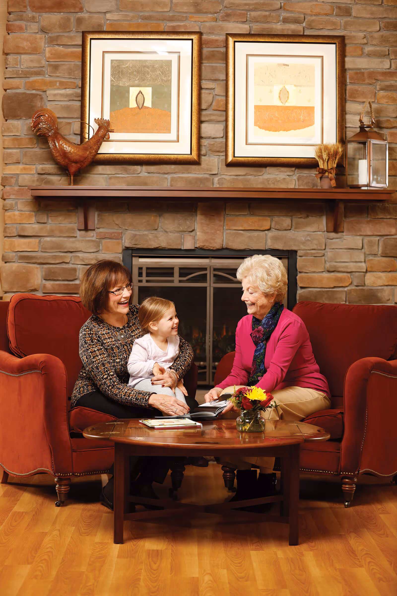 Three generations of women, a grandmother, mother, and young child, sitting on red armchairs around a wooden coffee table in front of a stone fireplace. They are smiling and looking at a book together. The room has wooden flooring, a mantel with decorative items, and two framed artworks above the fireplace.