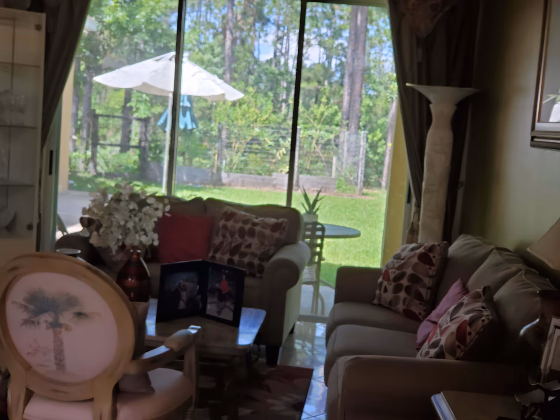 A cozy living room with two beige sofas adorned with patterned and solid color cushions, a wooden chair with a palm tree design on the backrest, a small wooden coffee table with a photo frame and a vase of white flowers. Large sliding glass doors reveal a green backyard with trees and a white patio umbrella.