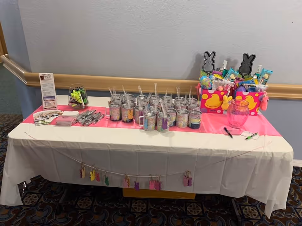 A table covered with a white tablecloth and a pink runner, displaying multiple clear plastic cups with straws and bunny designs, two colorful gift baskets filled with various items, a pink glass jar, pens, and informational pamphlets. The table is set against a light gray wall with a wooden chair rail and a patterned carpeted floor.