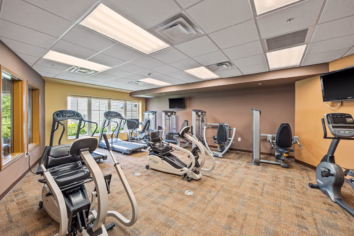 Well-lit fitness room with treadmills, recumbent bikes, and strength equipment arranged on a carpeted floor.