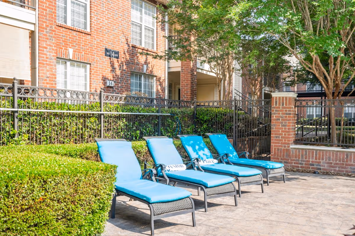 Turquoise poolside lounge chairs on a patio in front of a brick building, metal fence, and greenery.