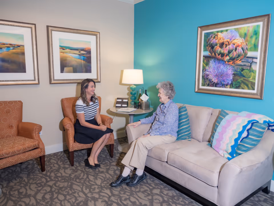 A cozy living room with a beige couch adorned with colorful pillows and a crocheted blanket. An elderly woman and a younger woman are sitting and smiling at each other. The room has two orange armchairs, a round side table with a lamp, and framed artwork on the walls, one wall painted teal.