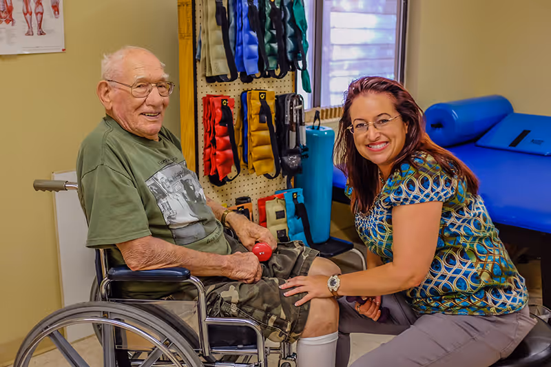 An elderly man in a wheelchair holding a small red dumbbell is assisted by a smiling female caregiver or therapist in a room equipped with various exercise and rehabilitation equipment, including weighted belts hanging on the wall and a blue therapy table.