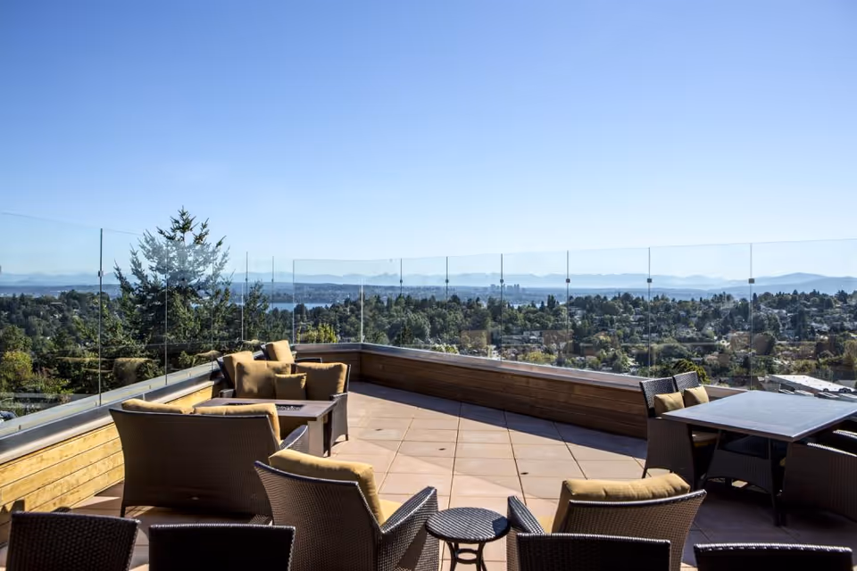 Outdoor patio area with cushioned wicker chairs and tables, surrounded by glass railing, overlooking a scenic view of trees, houses, and distant mountains under a clear blue sky.