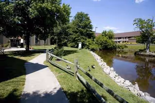 An outdoor path beside a pond with a wooden fence and trees near a campus-style building.