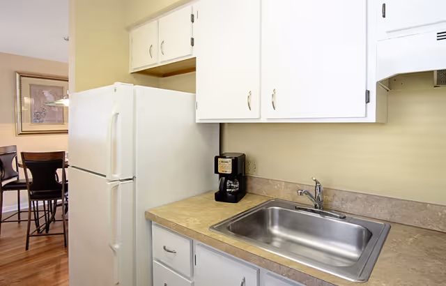 A small kitchen area with white cabinets, a stainless steel sink, a beige countertop, a white refrigerator, and a black coffee maker. In the background, there is a dining area with a table and chairs and a framed picture on the wall.