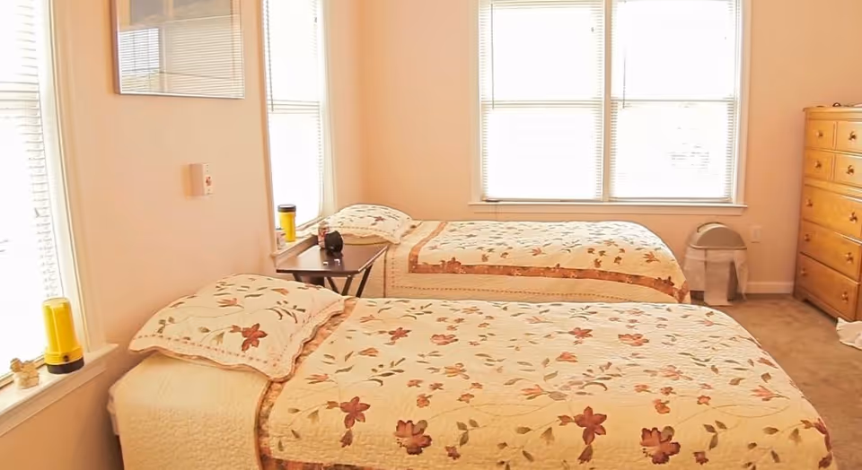 Sunlit bedroom with two twin beds covered in floral quilts, a small side table between them, and a wooden dresser.