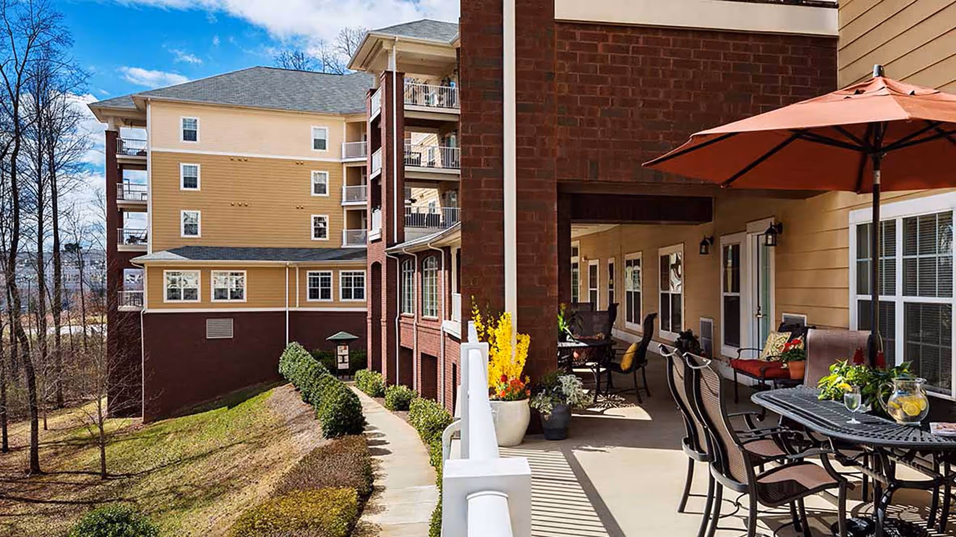 Outdoor patio area at Atria North Point with a table, chairs, an umbrella, potted plants, and a view of the building and surrounding landscape under a blue sky.
