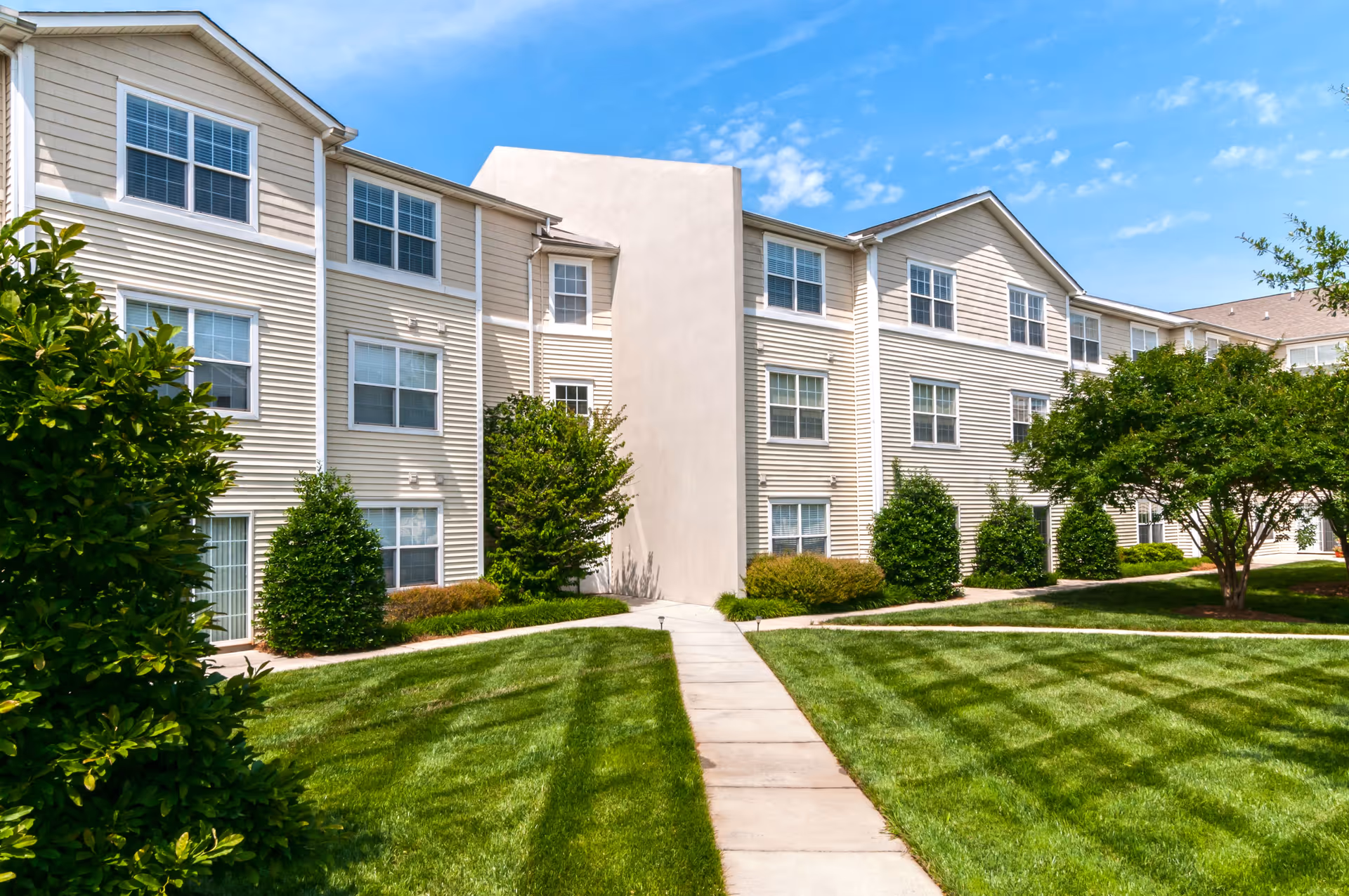 Exterior view of a three-story residential building with beige siding and multiple windows. A concrete walkway leads through a well-maintained green lawn with bushes and trees on either side under a blue sky with some clouds.