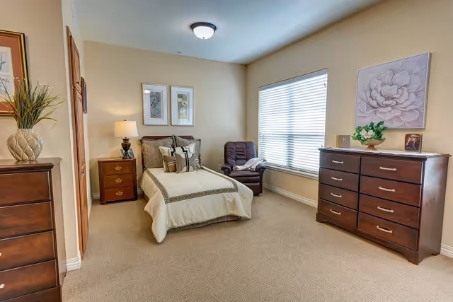 A cozy bedroom with a single bed dressed in beige and brown bedding, a wooden nightstand with a lamp, a comfortable armchair near a large window with blinds, and two wooden dressers. The walls are painted light beige and decorated with framed artwork and a flower painting.