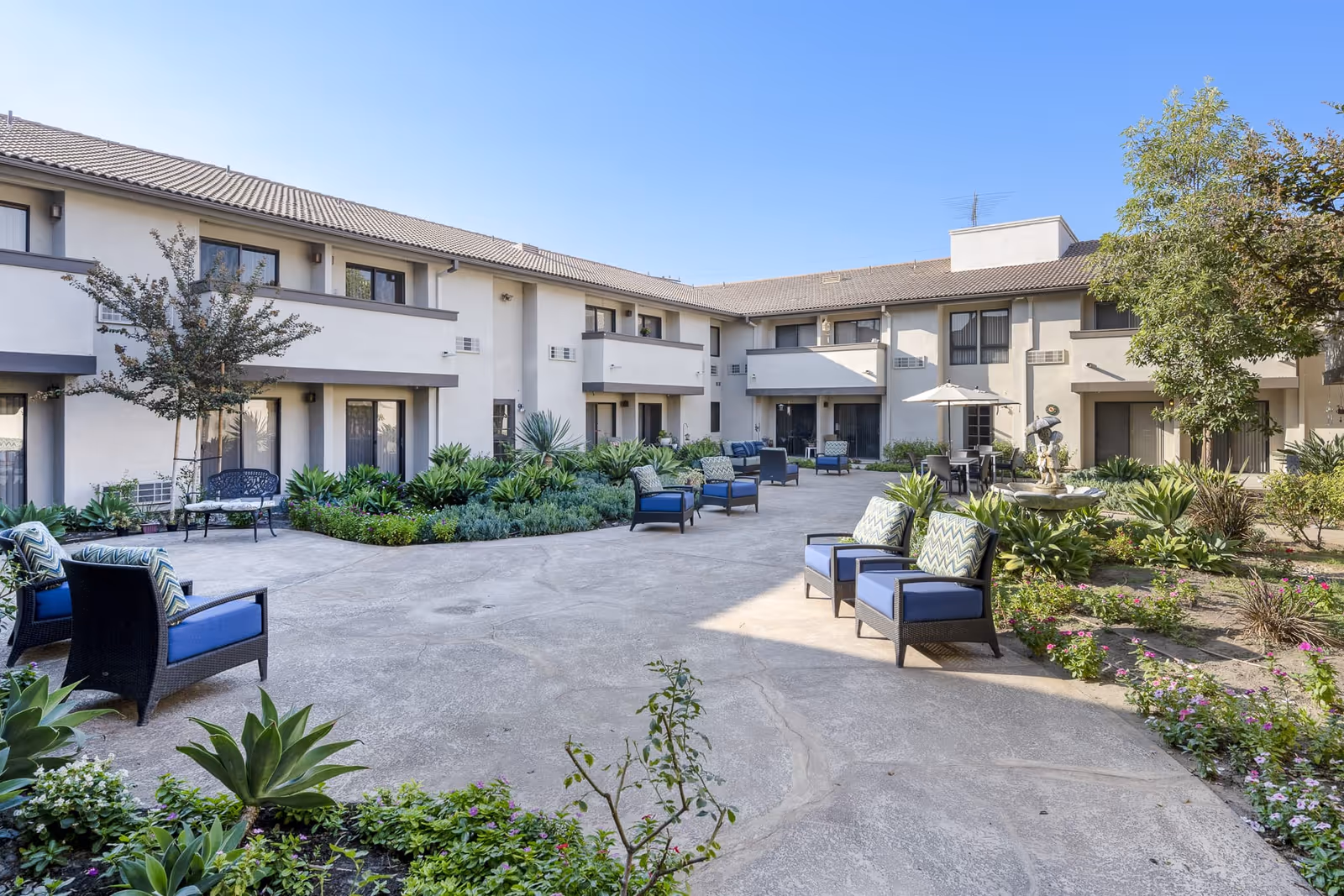 Sunlit courtyard of a senior living complex with outdoor seating, landscaped planters, and a two-story surrounding building.