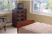 A small bedroom featuring a bed, a dark dresser with a small TV, and windows.