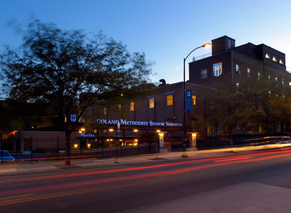 Dusk exterior view of a multi-story brick senior care facility with illuminated signage and streaking car lights in front.