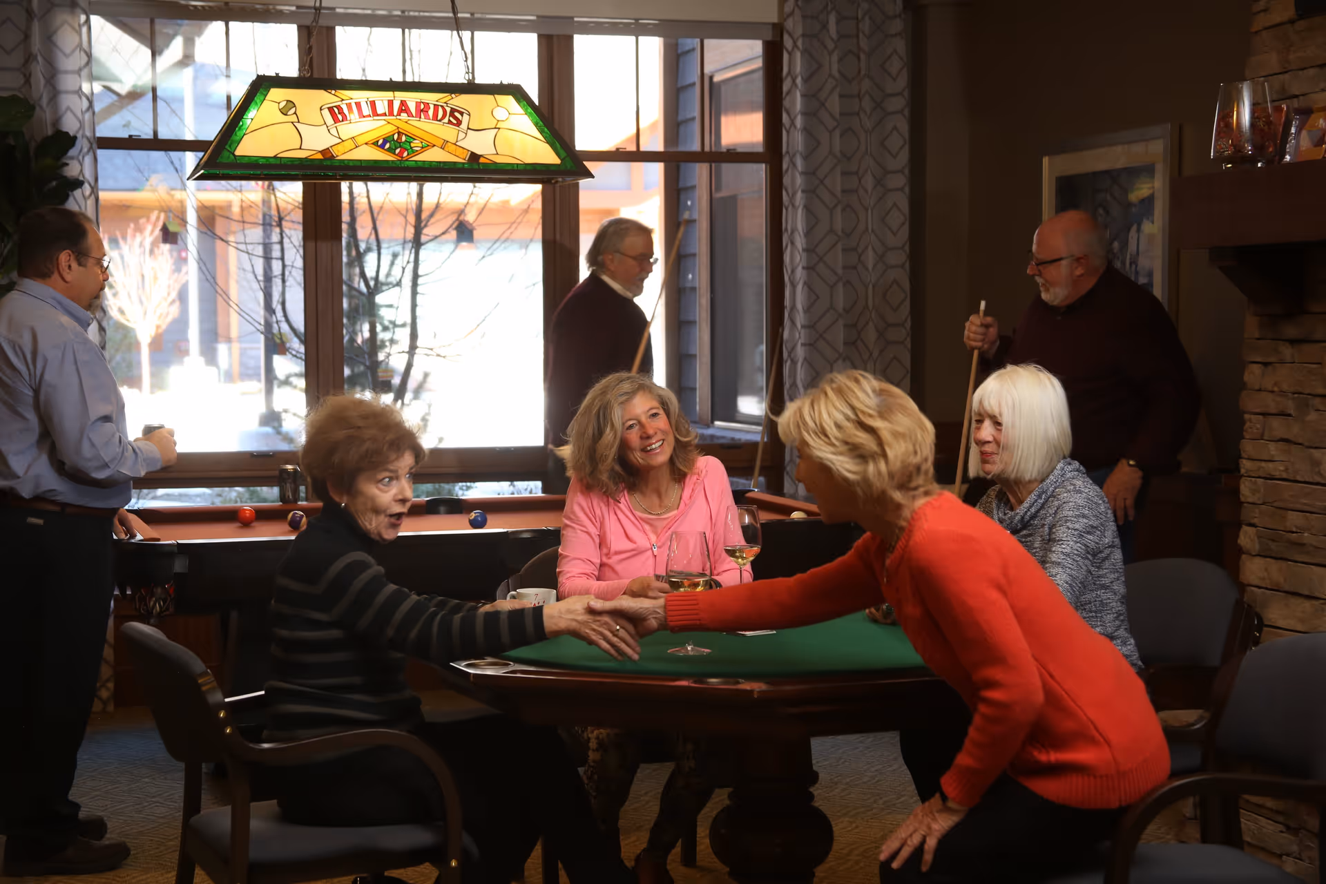 A group of elderly people socializing in a cozy indoor recreational room. Two women are shaking hands across a card table, while another woman and a man sit nearby. In the background, two men are playing billiards under a stained glass lamp that says 'BILLIARDS'. The room has large windows with curtains and a stone fireplace.