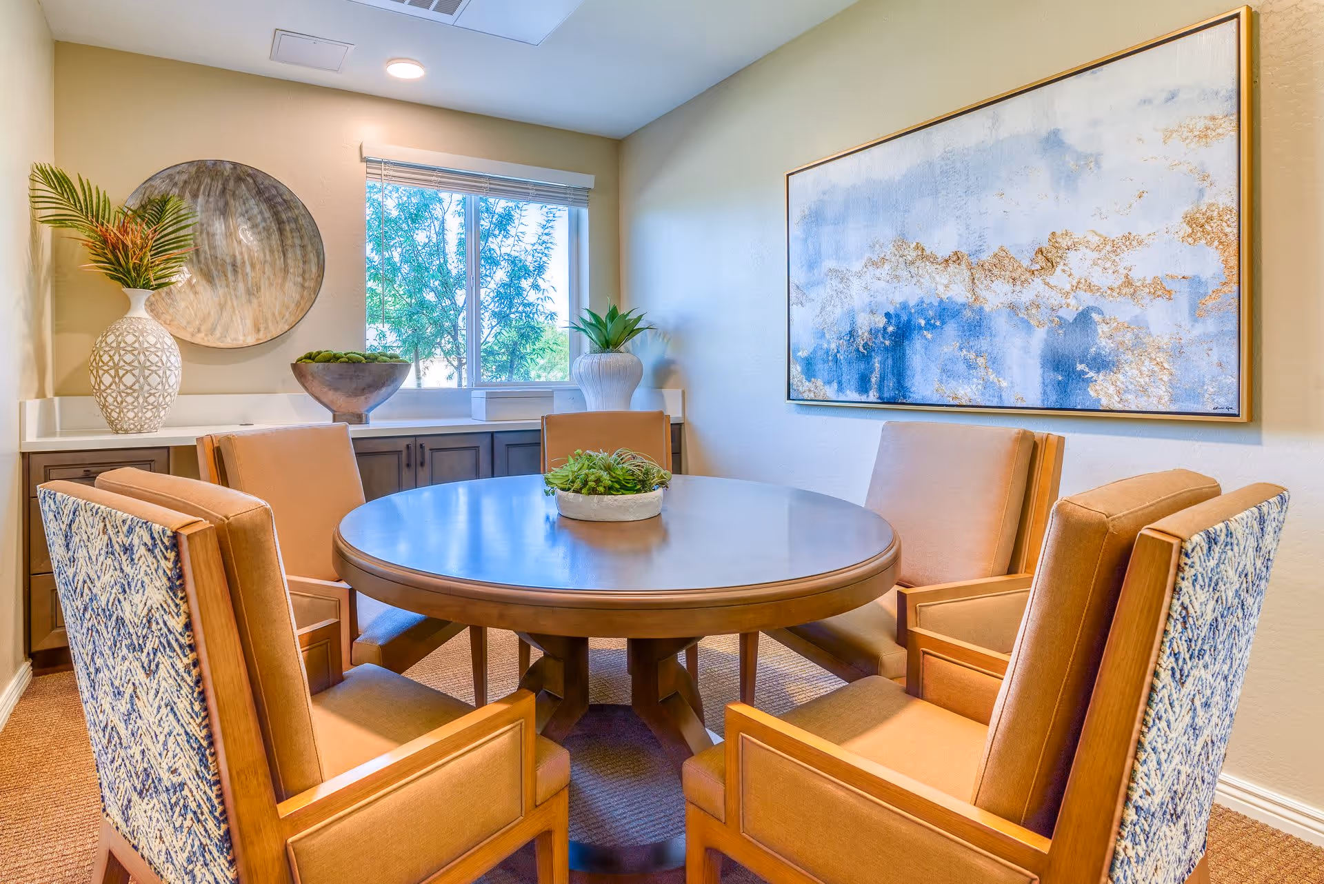 A bright dining room with a round wooden table surrounded by six cushioned chairs, two of which have patterned backs. The room features a window with a view of green trees, a sideboard with decorative vases and a bowl, and a large abstract painting with blue and gold tones on the wall.