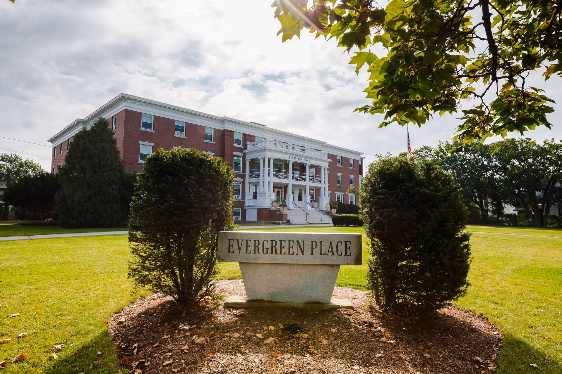 A large, three-story brick building with white columns and a porch labeled 'Evergreen Place' in the background, with a stone sign reading 'EVERGREEN PLACE' in the foreground surrounded by bushes and a well-maintained lawn under a partly cloudy sky.