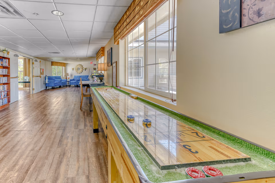 Long common area interior with a shuffleboard table along a window and seating with blue patterned chairs in the background.