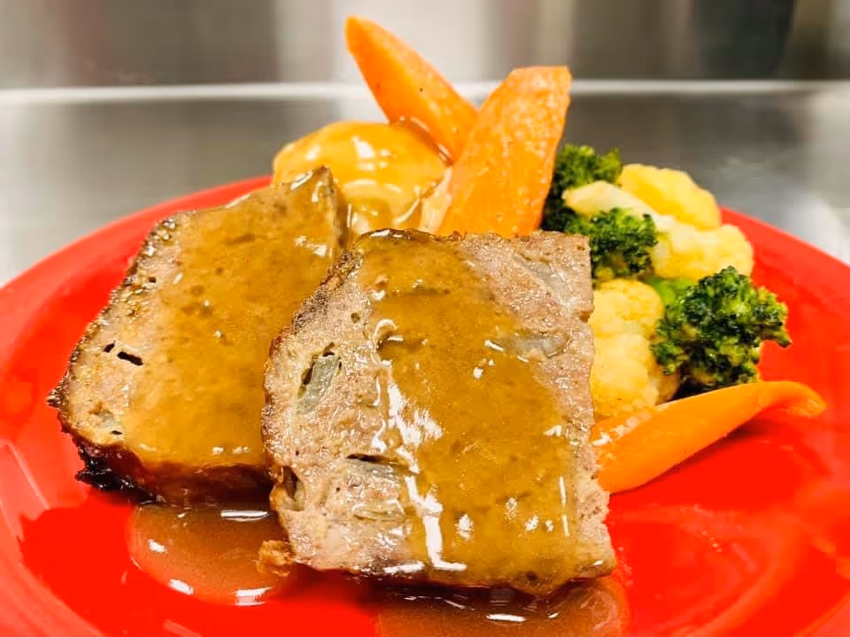 A red plate with two slices of meatloaf topped with brown gravy, served with cooked carrots, broccoli, cauliflower, and mashed potatoes with gravy in the background.