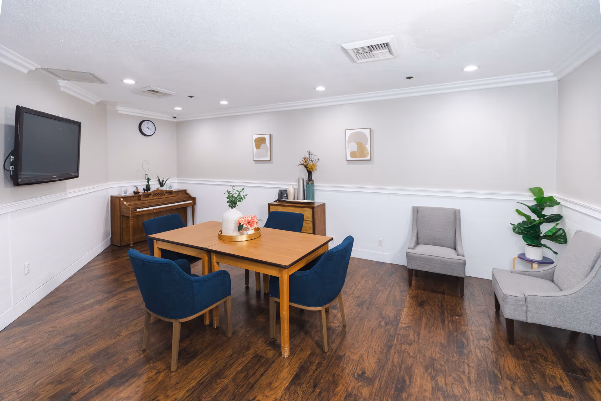 A small common room with wooden flooring and white walls featuring a wooden table with four blue chairs around it. There is a piano against the far wall with a clock above it, a flat-screen TV mounted on the left wall, and two gray armchairs with a small side table and a potted plant on the right. Two abstract art pieces hang on the wall above a small wooden cabinet with decorative items.