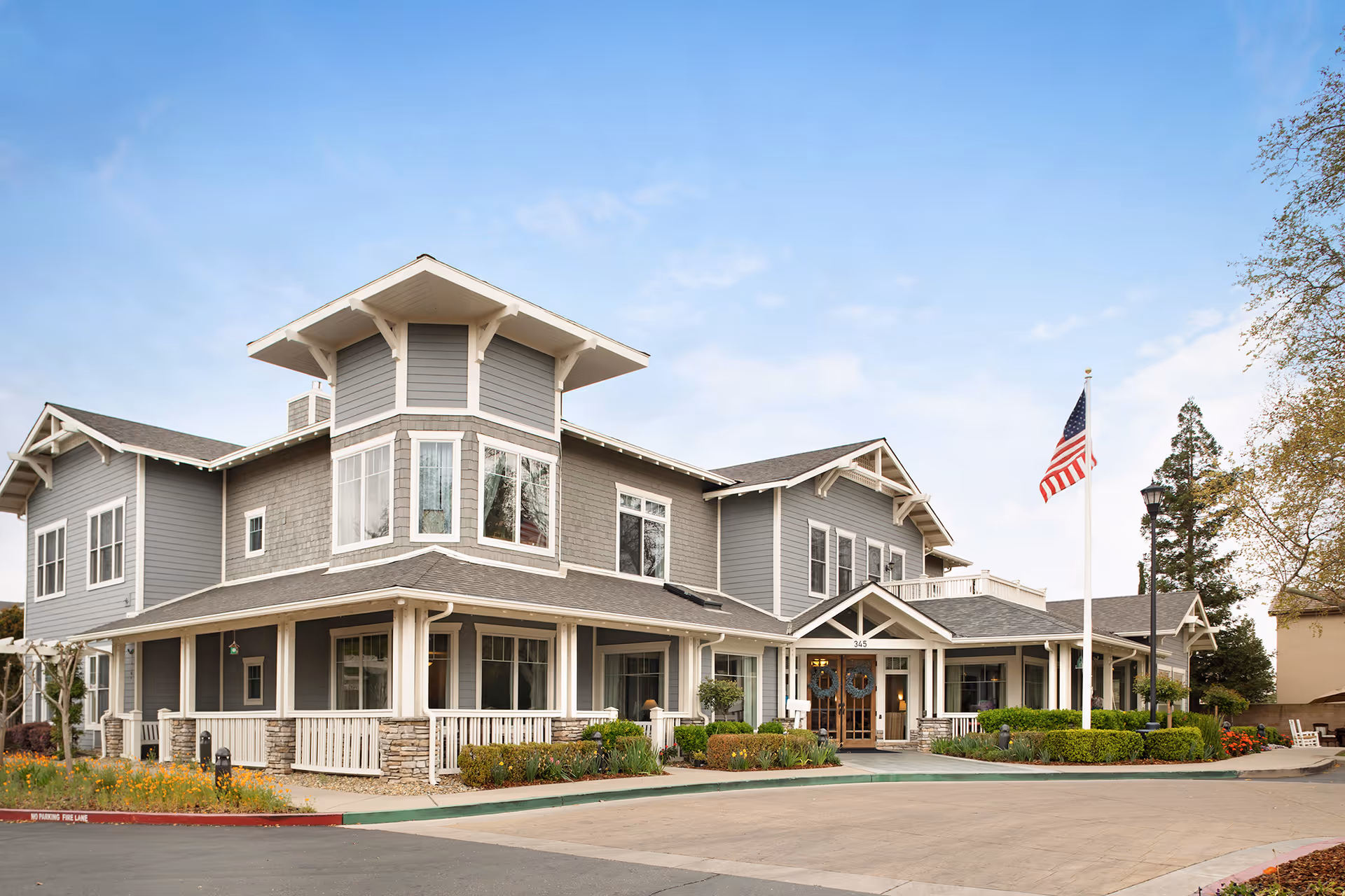 Front exterior of a two-story gray senior living building with a wraparound porch, landscaped entrance, and an American flag.