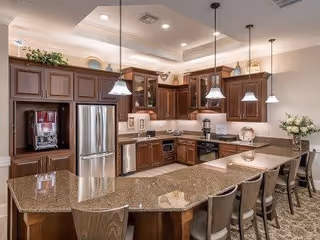 Spacious kitchen with dark wooden cabinets, granite countertops, and stainless steel appliances including a refrigerator and oven. The kitchen features a large island with seating for five, pendant lighting, and decorative plants and dishes on top of the cabinets.