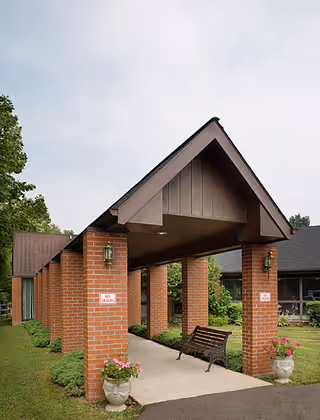 Covered brick portico entrance with a peaked roof, bench, and potted flowers leading to the building.