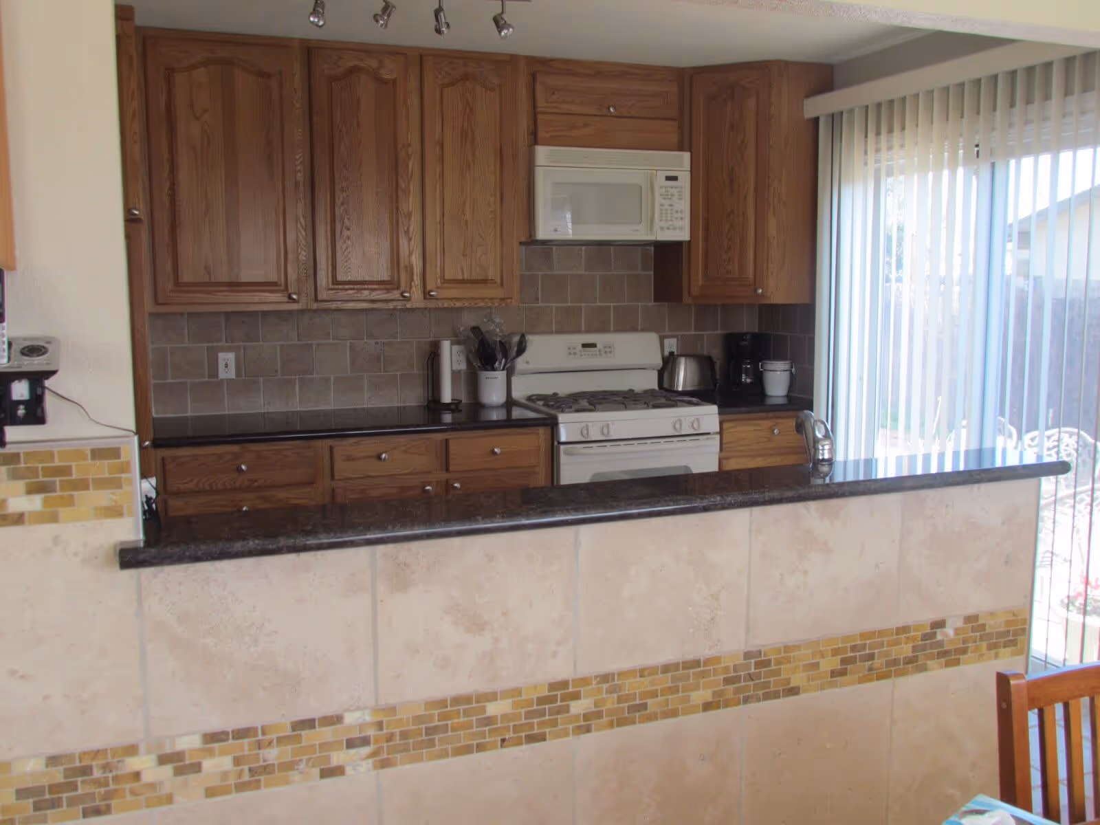 Kitchen with oak cabinets, a white stove and microwave, a dark granite countertop and tiled breakfast bar by a sliding glass door.