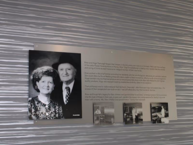 Wall-mounted commemorative plaque with a large black-and-white portrait of an elderly couple, accompanying text and three small photos on a textured wall.