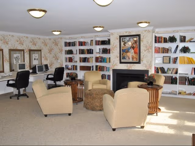 A cozy senior community room with four beige armchairs arranged around a round ottoman and two small wooden side tables with plants. The room features built-in white bookshelves filled with books and decorative items, a fireplace with a framed artwork above it, and a computer workstation area with two desktop computers and black office chairs. The walls have floral wallpaper, and the room is well-lit with ceiling lights.