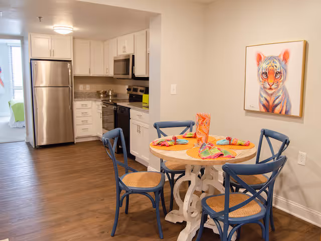 A cozy dining area with a round wooden table set for four with colorful napkins and a decorative vase. The table is surrounded by four blue chairs with woven seats. In the background, there is a modern kitchen with white cabinets, stainless steel refrigerator, microwave, stove, and granite countertops. A framed painting of a tiger cub hangs on the wall to the right.