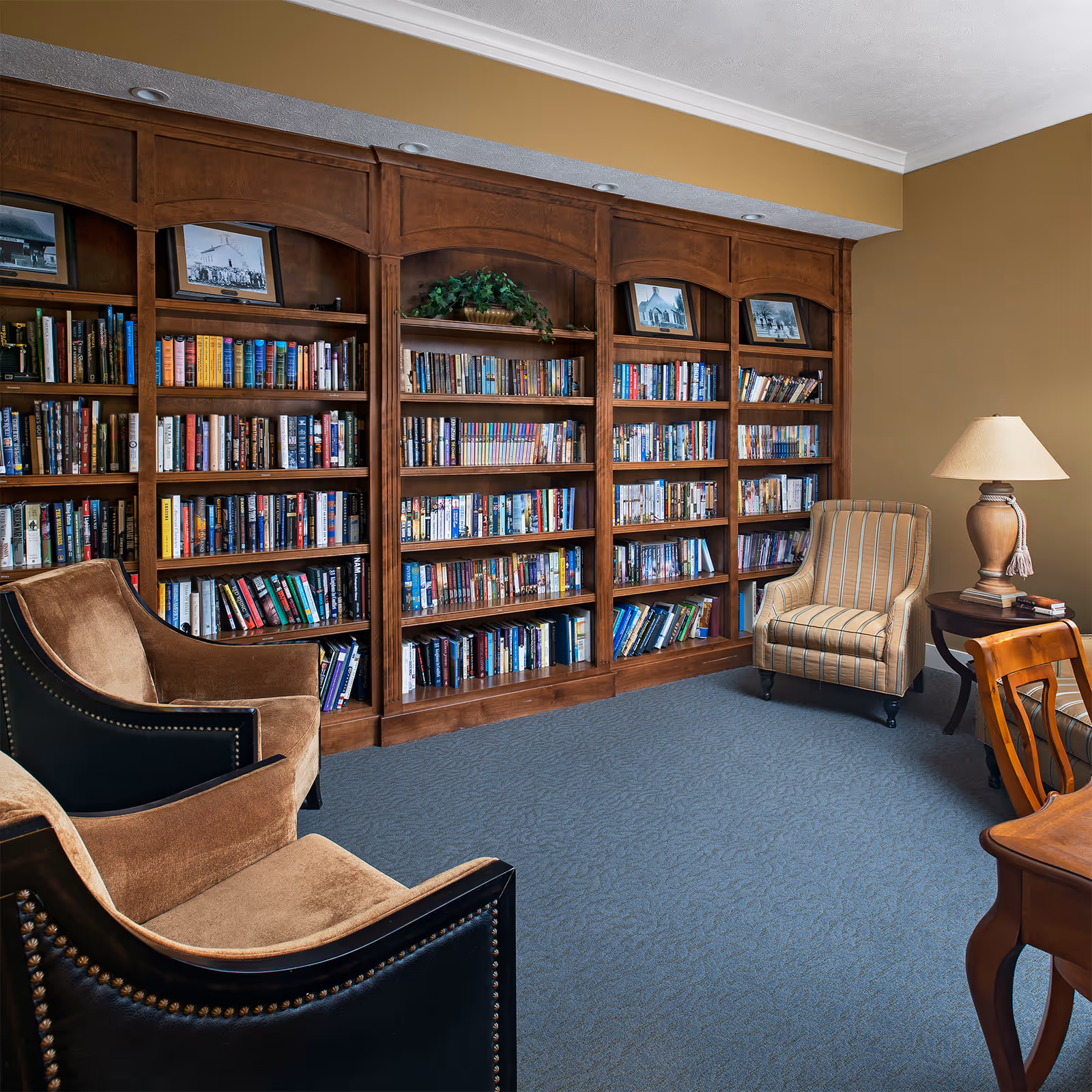 Cozy reading room with a large wooden bookcase filled with books, upholstered chairs, a side table and lamp.