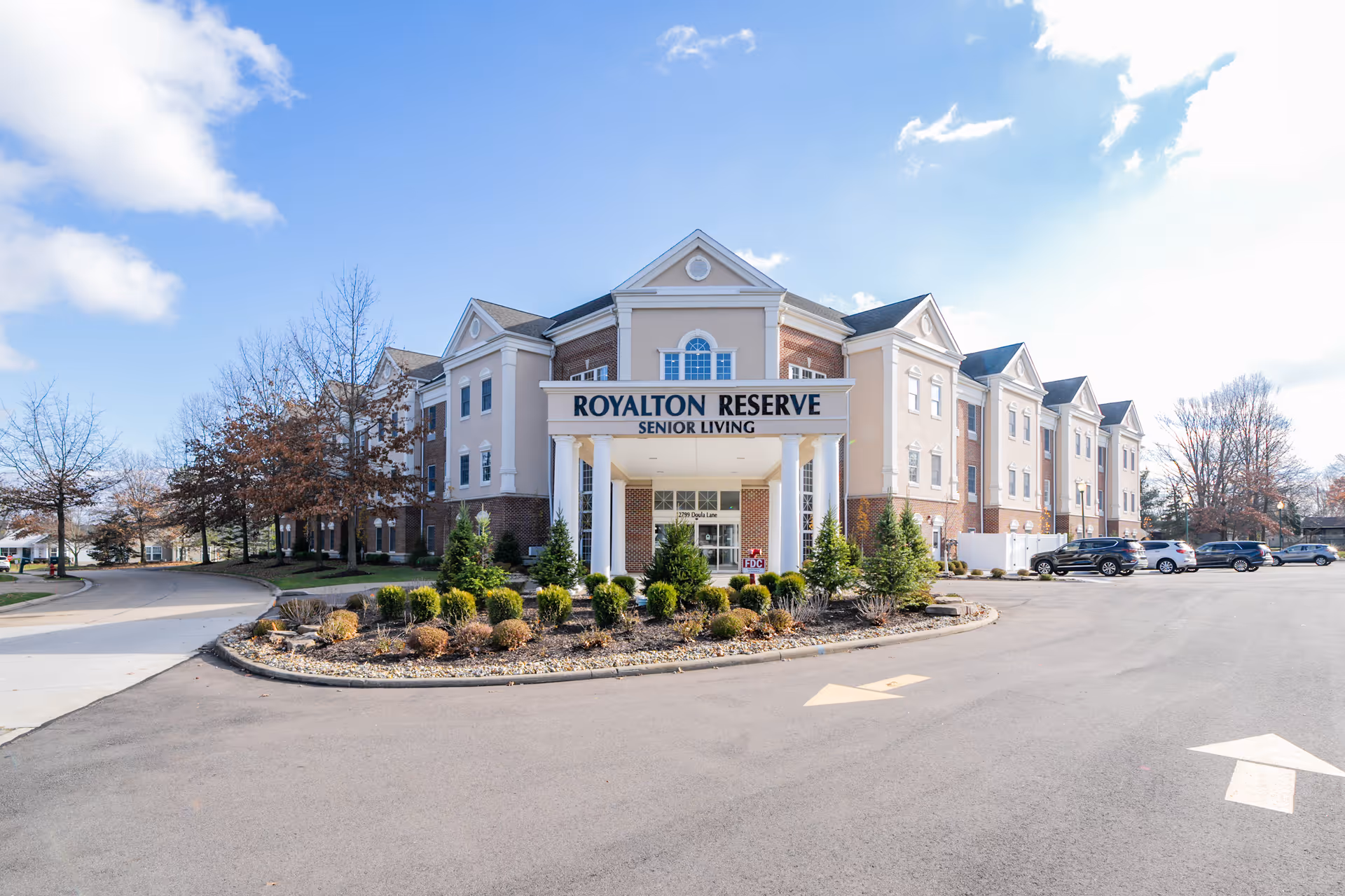 Exterior view of Royalton Reserve Senior Living facility, a multi-story building with a covered entrance supported by white columns, surrounded by landscaped bushes and trees, with a parking lot and several cars visible under a partly cloudy sky.