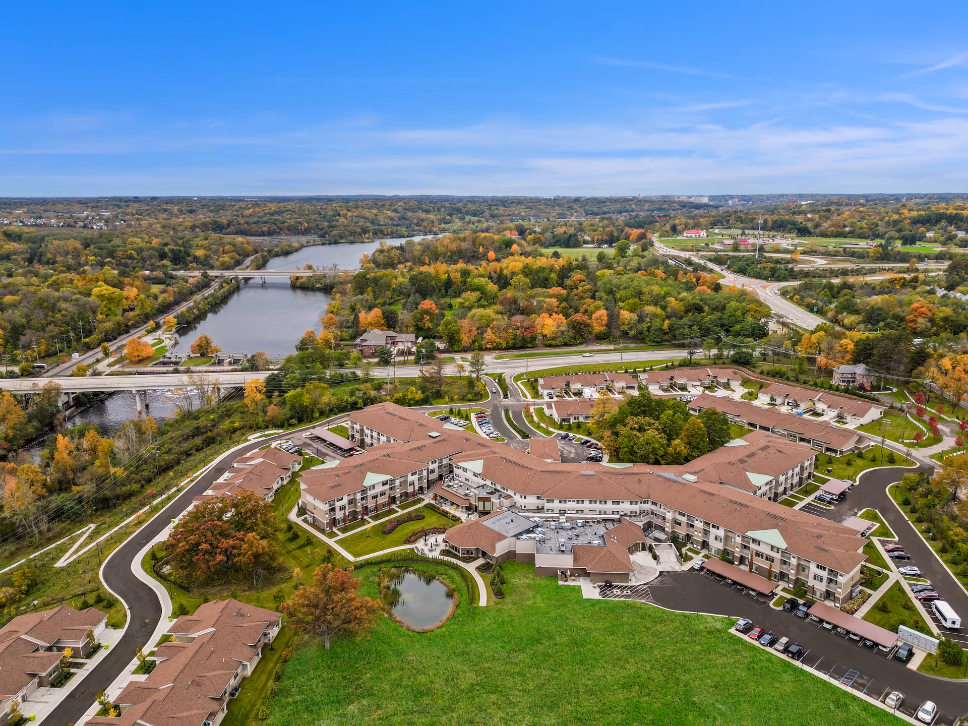 Aerial view of a large senior living facility named All Seasons Ann Arbor surrounded by green lawns, trees with autumn foliage, a small pond, and nearby roads and bridges over a river.