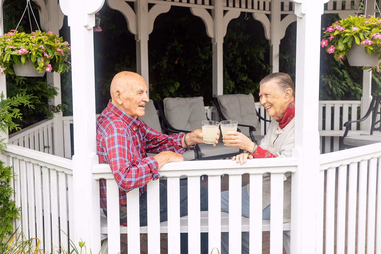 Two elderly people sitting in a white gazebo, smiling and clinking glasses filled with a light-colored beverage, surrounded by hanging flower pots and greenery.