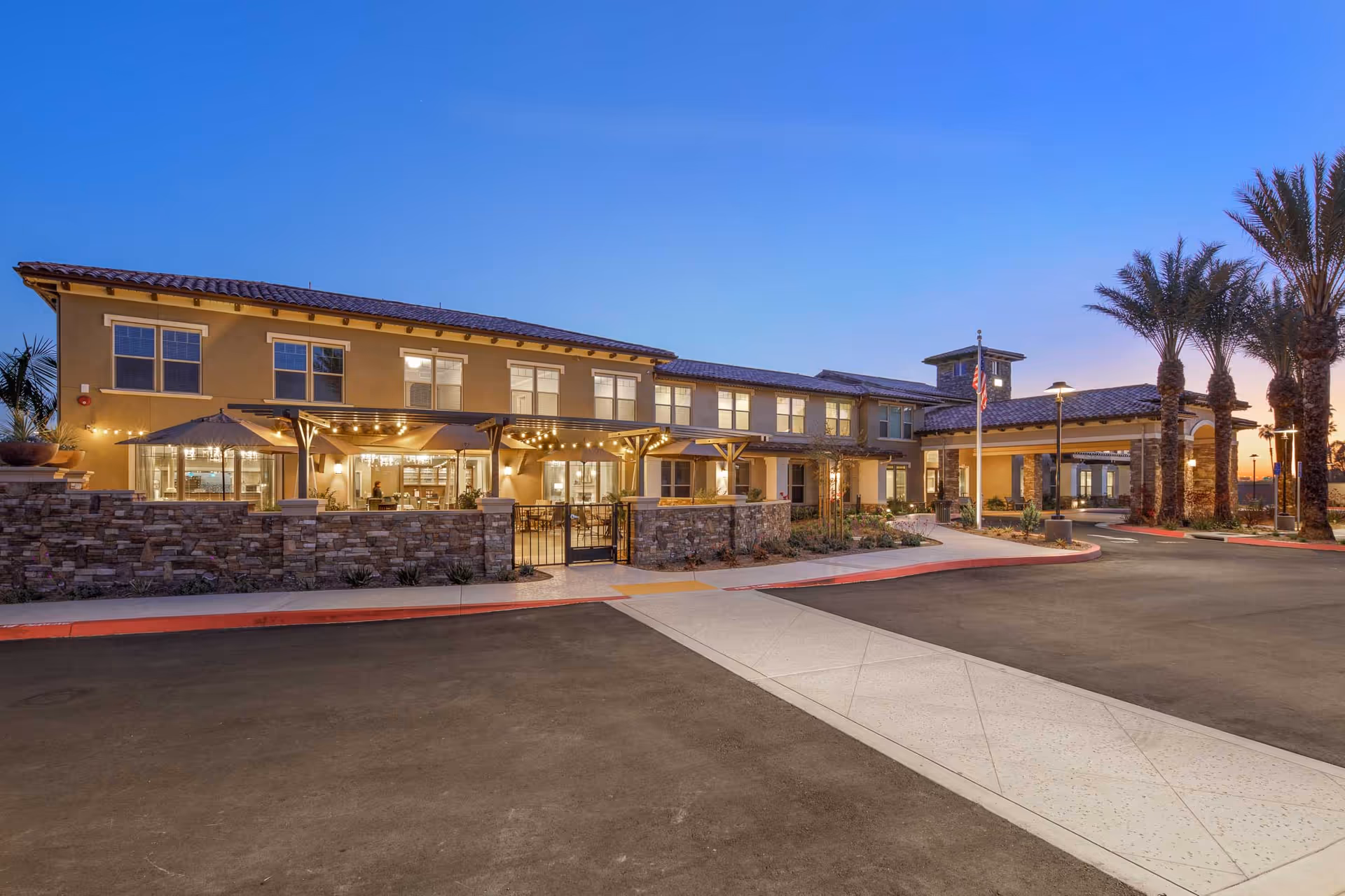 Exterior view of Oakmont of Riverpark senior living facility at dusk, showing a two-story building with warm lighting, stone accents, a covered entrance, palm trees, and a paved driveway.
