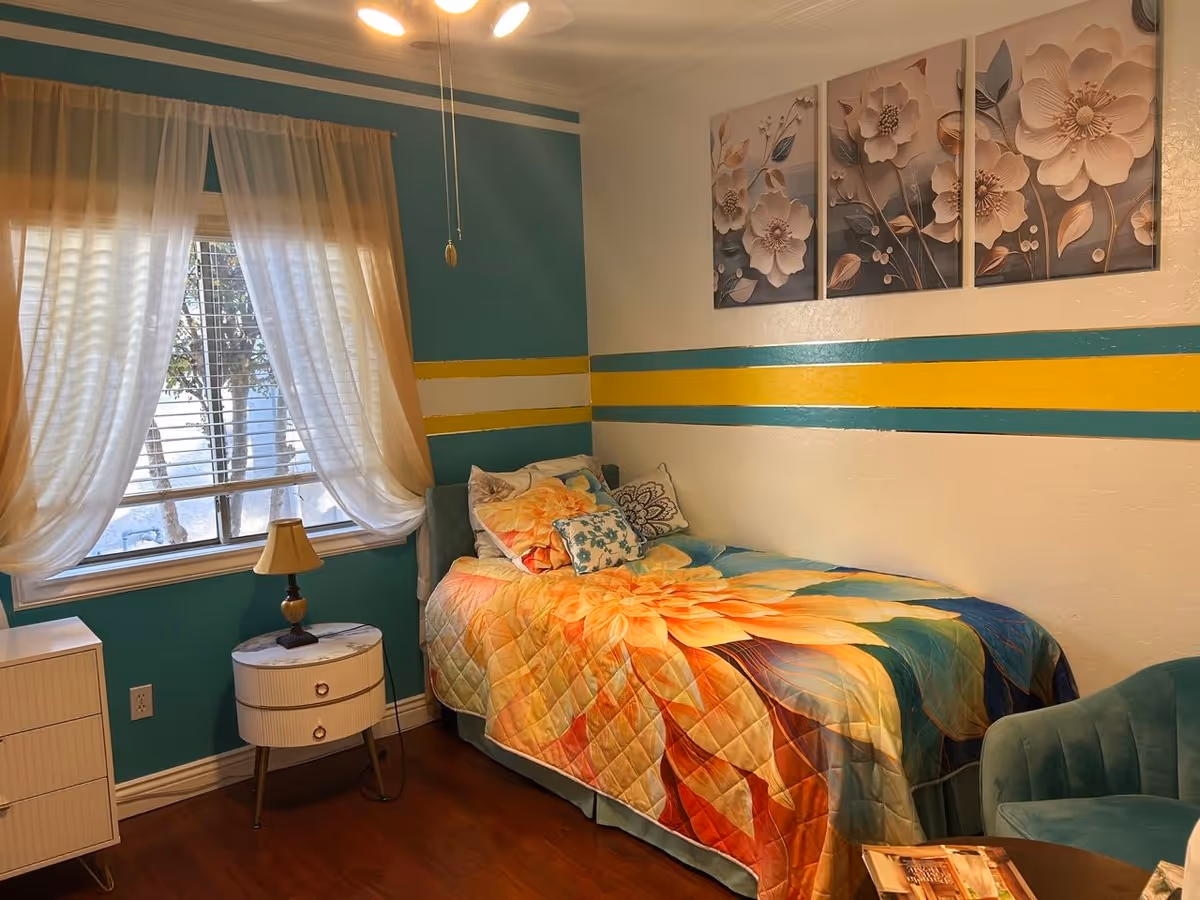 A cozy bedroom with a single bed covered in a colorful floral quilt and multiple pillows. The walls are painted teal and white with yellow and teal stripes. There is a window with sheer curtains letting in natural light, a small round bedside table with a lamp, a green upholstered chair, and three floral paintings hanging on the wall above the bed.